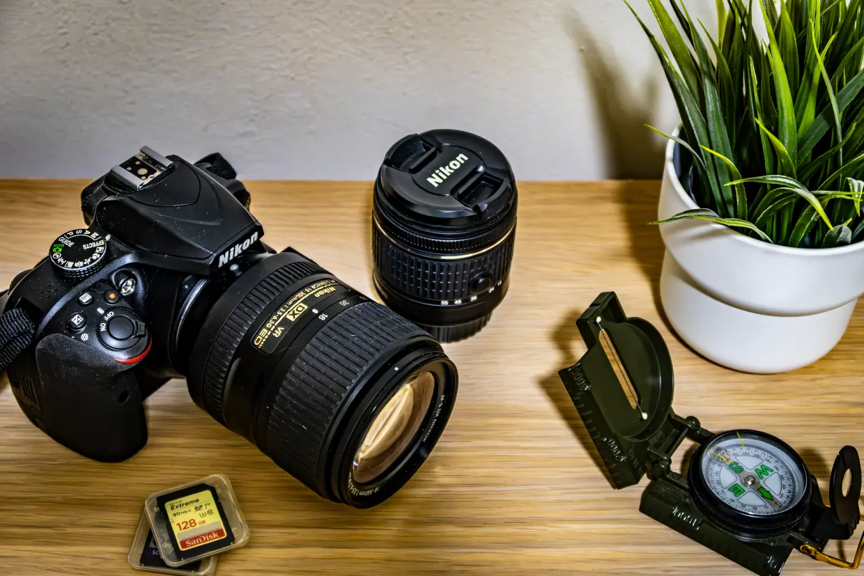 Photo of a Nikon D3400 camera with Nikon AF-S DX Nikkor 18-300mm f/3.5-6.3G ED VR lens mounted, with a couple of SD cards around, an open dark green metal compass, and a fake green plant on a white pot, all o them over a table.